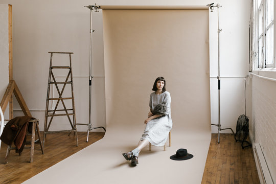 Behind The Scenes Of Model Sitting In Front Of Backdrop Seamless Paper In Photo Studio With Props Around