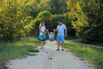 Fototapeta premium Parents swing the little boy in their arms like a swing and walk along the path in the Park at the sunset of a summer day. The concept of family happiness. Shut. Horizontal view