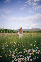 A young girl in a hat stands in a daisy field among flowers and spikelets. The concept of summer days and traveling