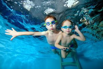 Two cheerful sports little boys swim in the pool, on a blue background. View from the bottom of the pool. Pose for the camera with your eyes open. Portrait. Underwater photography. Horizontal view