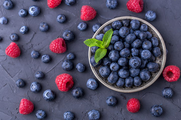 ripe, large blueberries in a metal plate, with a leaf of mint, scattered berries around, raspberries. Dark grey stone table