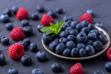 ripe, large blueberries in a metal plate, with a leaf of mint, scattered berries around, raspberries. Dark grey stone table
