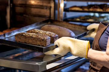 young baker on rubber gloves holding a tray with fresh bread taking them out of oven.