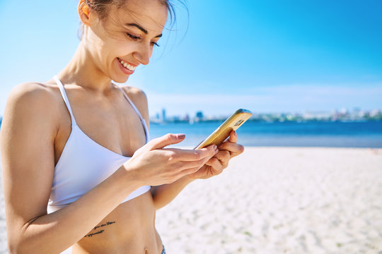 Closeup portrait of beautiful sexy happy girl on the sand city beach with sea background. Young smiling woman standing with the smartphone in the hand, looking at phone against blue sky and sea