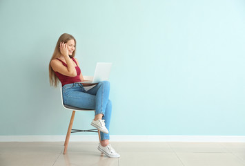 Beautiful young woman with laptop sitting on chair against color wall