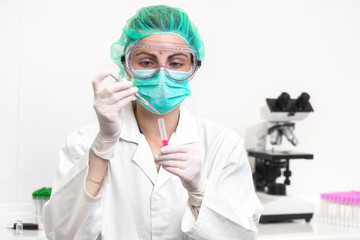 Young attractive concentrated female scientist in protective eyeglasses, mask and gloves dropping a red liquid substance into the test tube with a pipette in the laboratory .