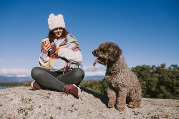 A young beautiful woman is sitting with her spanish water dog on the stone in the nature. She is looking at her dog that is sitting next to her. The dog is sitting while looking her owner.