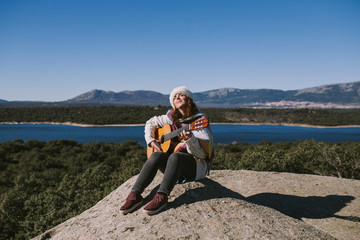 Fototapeta premium A beautiful young woman is sitting on the stone while playing the guitar. Behind her there is a beautiful landscape of mountains and a big lake. She is alone in the middle of the nature.