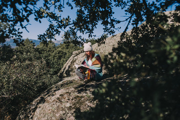 Obraz premium A young woman is sitting on the stone while reading a book. She is in the middle of the forest enjoying the nature. She is wearing a wool hat and a colorful foulard. It is a beautiful sunny day.