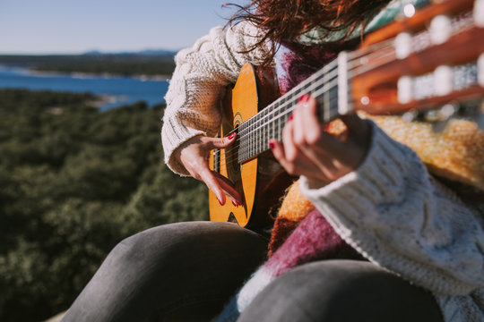 Close up. A young woman is playing the spanish guitar in the nature. Her fingers are touching the strings of the guitar. Her nails painted in red. She is wearing a white jacket and a colorful foulard.