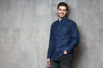 Obraz premium European male leaning on gray wall in denim shirt, looking confidently at camera