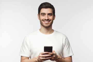 Obraz na płótnie Canvas Young handsome man holding smartphone and looking at camera with smile while standing isolated on gray background