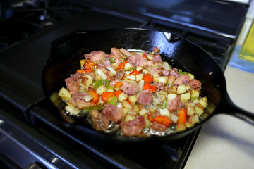 Corned beef hash frying in a cast iron skillet on a natural gas stove top in a home kitchen.