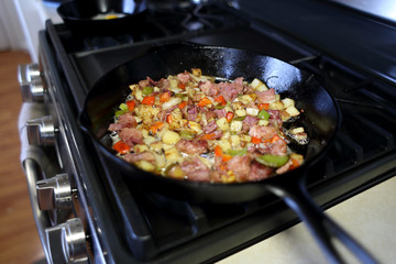 Corned beef hash frying in a cast iron skillet on a natural gas stove top in a home kitchen.