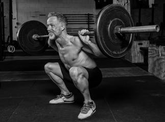 Ein durchtrainierter Mann mittleren Alters macht oberkörperfrei Squats. Fitnesstraining mit der...