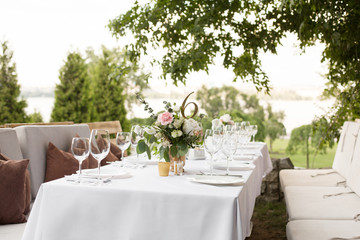 Wedding table setting decorated with fresh flowers in a brass vase. Wedding floristry. Banquet table for guests outdoors with a view of green nature. Bouquet with roses, eustoma and eucalyptus leaves