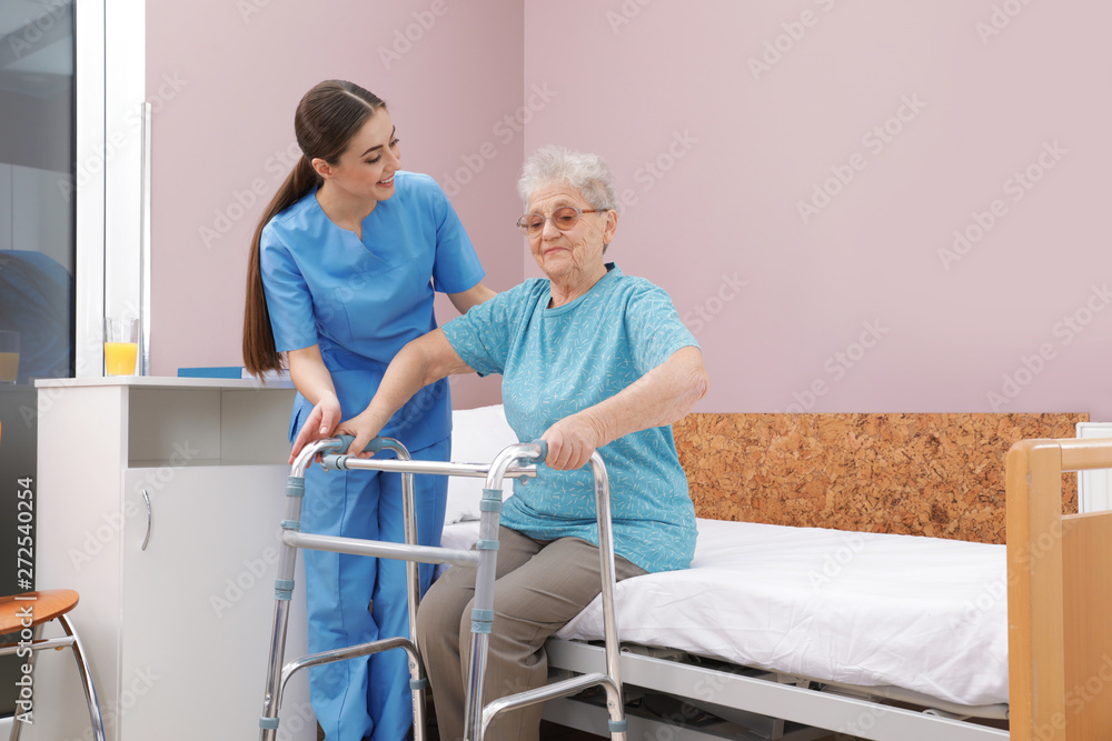 Sticker nurse assisting senior woman with walker to get up from bed in hospital ward