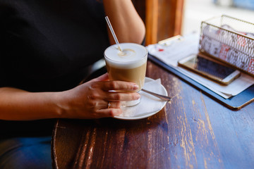 Young girl drinks a hot latte from a large glass at a wooden table located by the window in a cozy cafe. Woman's hands and freshly brewed coffee breakfast at the wooden table