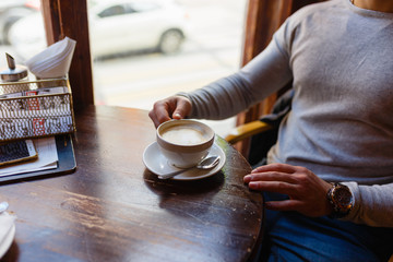 Man drinks a hot cappuccino from a large white mug at a wooden table located by the window in a cozy cafe. Men's hands and freshly brewed coffee breakfast at the wooden table