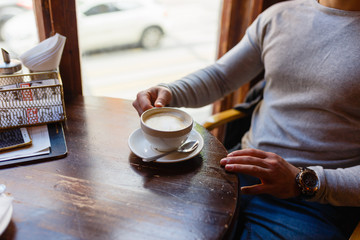 Fototapeta na wymiar Man drinks a hot cappuccino from a large white mug at a wooden table located by the window in a cozy cafe. Men's hands and freshly brewed coffee breakfast at the wooden table