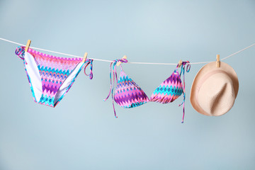 Stylish bikini and hat hanging on rope against color background