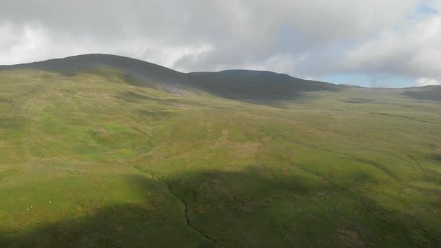 An aerial backward footage of a majestic mountain summit grassy slope with a trail path, blue sky and huge altitude white clouds