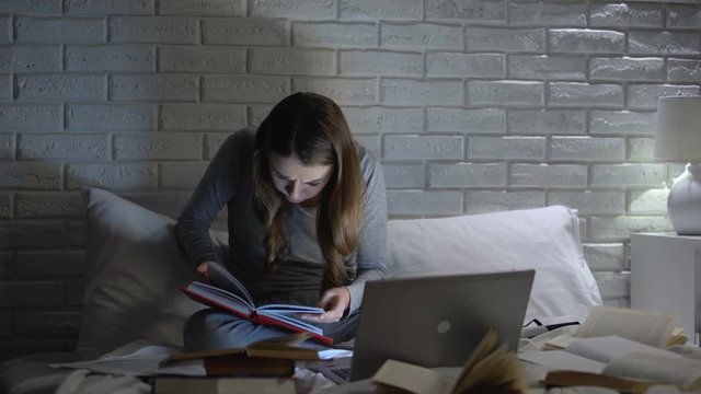 Female student reading books, typing project on laptop late at night, deadline