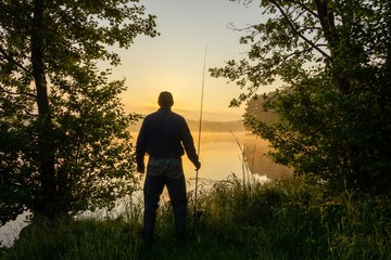 Silhouette of angler standing on the lake shore during misty sunrise