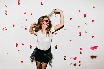 Elegant young lady in trendy sunglasses looking up at fallen hearts. Portrait of positive long-haired woman in short skirt enjoying photoshoot.