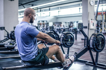 Male using rowing machine at fitness club. Young man doing exercises on fitness machine in gym. Side view - obrazy, fototapety, plakaty