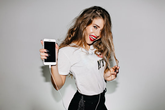 Charming caucasian girl with smartphone in hand having fun during indoor photoshoot. Photo of ecstatic curly lady in stylish t-shirt listening favorite music isolated in white background.