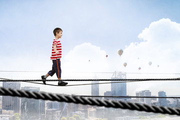 Cute joyful little boy walking on a rope over city. Mixed media