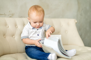 Fototapeta premium Happy chubby blond boy sitting on a light sofa and looking at a book. A red-haired boy in blue jeans and a white shirt is looking at a closed book. Horizontal photography