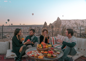 Four friends or two couples have fun whilst having breakfast outdoor with fruits, breads and drinks with a beautiful view to the balloons in Cappadocia, Turkey 