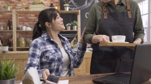 Friendly waiter server laughing smiling having fun with customer patron in modern coffee shop. young asian japanese waitress giving beverage client order and sharing discussing with friend cellphone