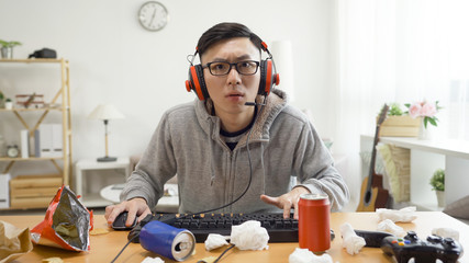 Man addicted to computer games. concentrated young asian male in headset sitting in dirty apartment...