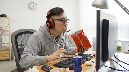 Teenager boy high school student looks at the computer screen holds chips in hand and plays video...