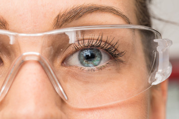 A beautiful young Caucasian is viewed closeup in the workplace, wearing protective goggles over her eyes. Pretty eyelashes and blue eyelashes seen in detail behind plastic eyewear. - obrazy, fototapety, plakaty
