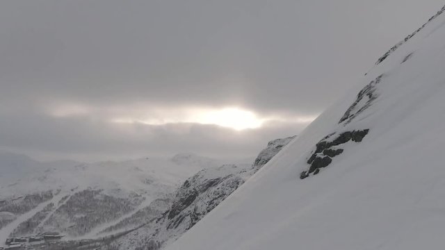 Snowy mountain side meets ground covered in snow. Cloudy day during snowfall. Pan and tilt to show the sun shining through clouds. Ends with snowy landscape and clouds. Clip H2 3 of 3