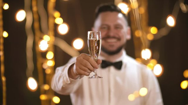 Portrait with selective focus of happy bearded man in white shirt and bowtie standing in room decorated with festive fairy lights and raising his champagne glass