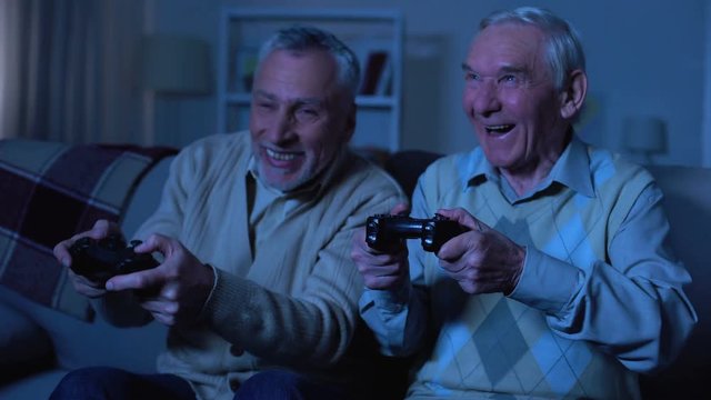 Emotional elderly male friends holding joysticks and playing video game, fun