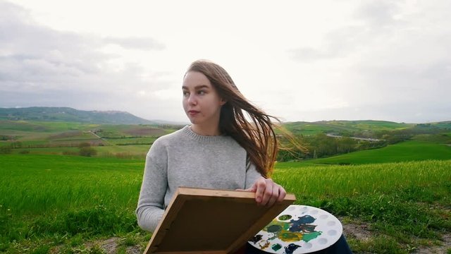 A young attractive woman sits in the middle of a green field and draw. Hair blown in the wind