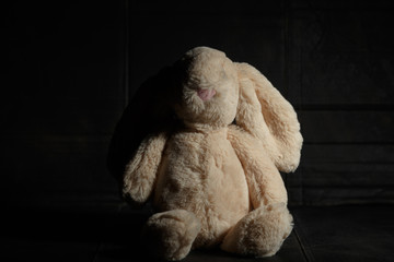 Soft toy bunny on a dirty dark background in the beam of hard light with shadow. Lost childhood...