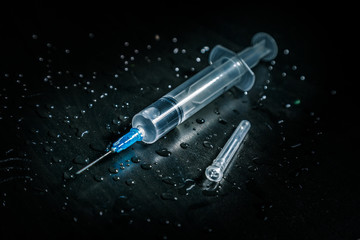 A syringe with a clear liquid lies on a dirty dark background covered with drops.