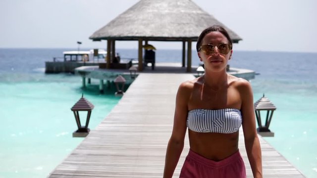 Woman in Bikini and Shorts Walks Pier Over Clear Aqua Water - Vabbinfaru, Maldives