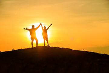 The couple stands holding hands on the top of the mountain with beautiful sunsets.happy man and woman tourist at hill,Happy couple with raised arms stands