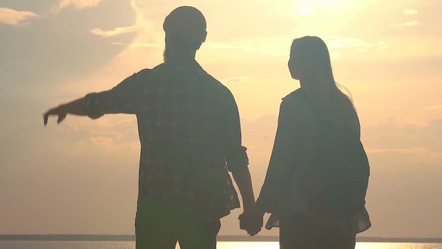 Silhouttes of young couple in love standing near the sea. They are holding hands, hugging, enjoying beatiful sunset over the sea.