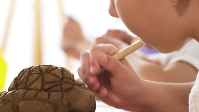 Creating toy made of clay in the workshop. Child is scratching with spicial tool the texture of the scales on the handmade turtle toy's head from clay, hand closeup.