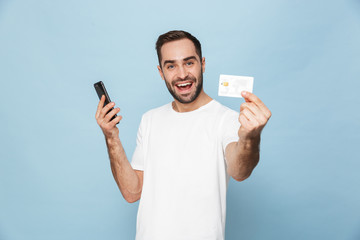 Photo of young caucasian man in casual white t-shirt rejoicing while holding credit card and...