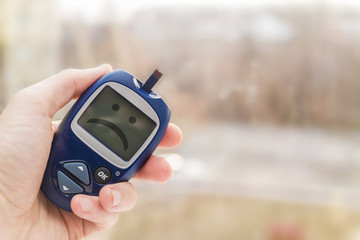 Man holding glucometer with test strip with sad smile on the monitor on blurred background. Blood glucose meter. Closeup, selective focus